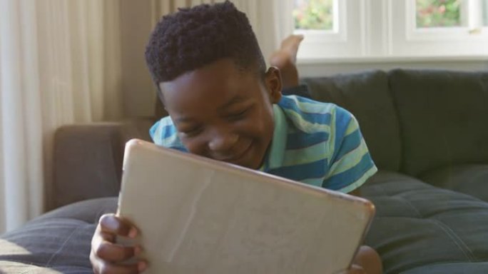 快乐的非洲裔美国男孩躺在沙发上，用平板电脑微笑