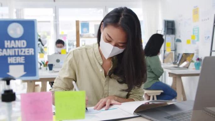 亚洲女商人企业家戴着医用口罩，在新的正常情况下保持社交距离，以预防病毒，同时在办公室工作时使用笔记本