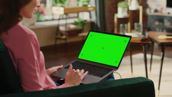 年轻的成年女性在带绿屏模拟显示的笔记本电脑上的家庭阁楼公寓工作。使用触控板和浏览互联网的创意女性。特