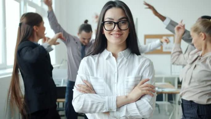 微笑的亚洲女孩站在办公室，双臂交叉，同事在背景中跳舞
