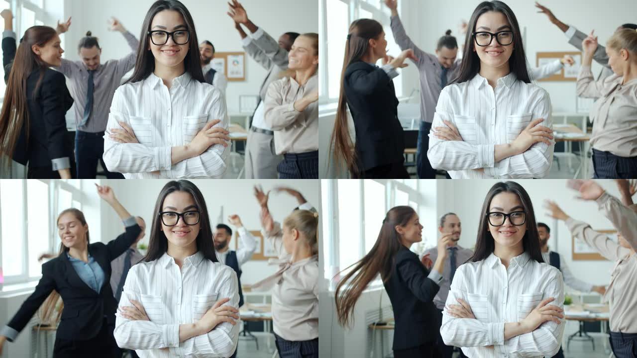 微笑的亚洲女孩站在办公室，双臂交叉，同事在背景中跳舞