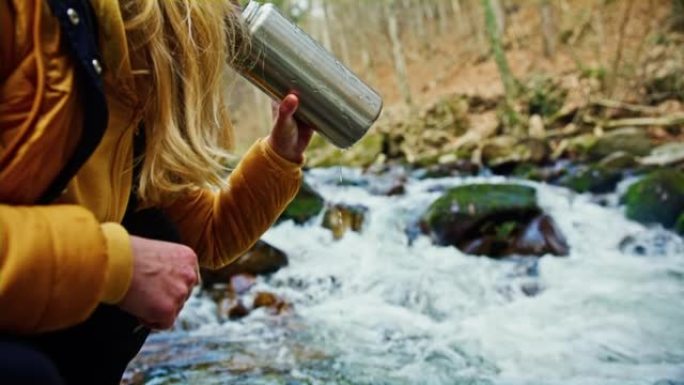 女人从森林溪流旁的热水瓶中填充水并喝水
