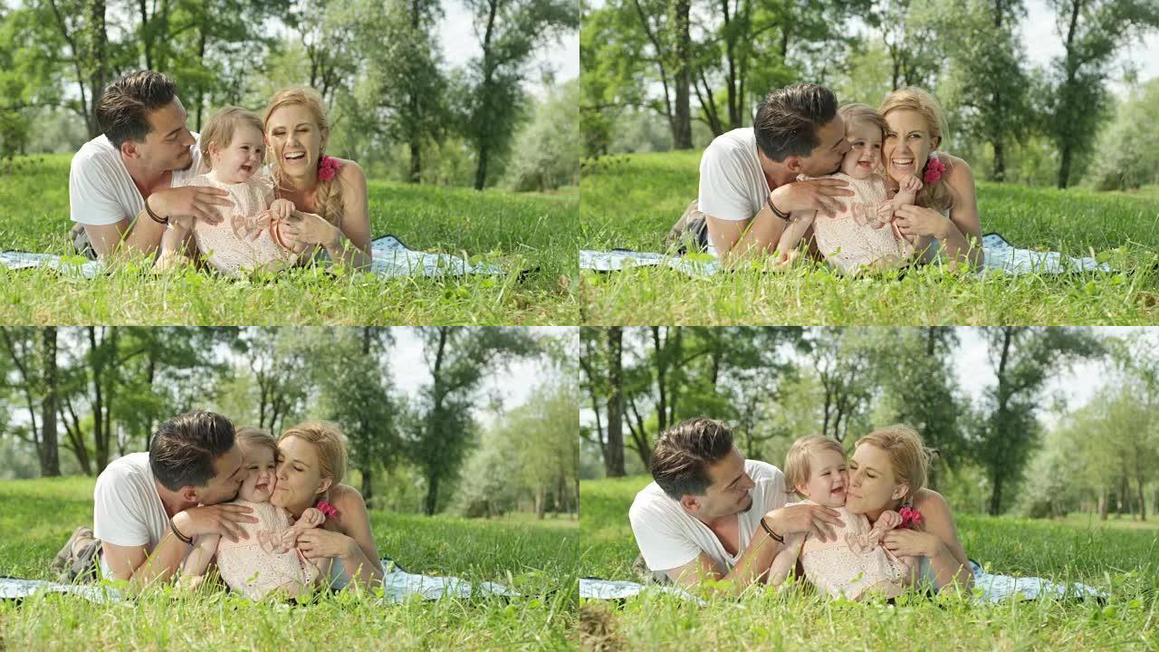 慢动作: 年轻的爸爸妈妈躺在公园的毯子上，亲吻宝贝女儿