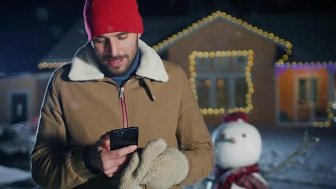 英俊的年轻人站在圣诞节前夕用花环装饰的田园诗般的房子的后院时使用智能手机。Guy使用手机向家人和朋友