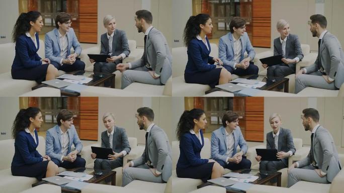 拥有数字平板电脑的女商人与坐在现代办公室沙发上的男女同事交谈