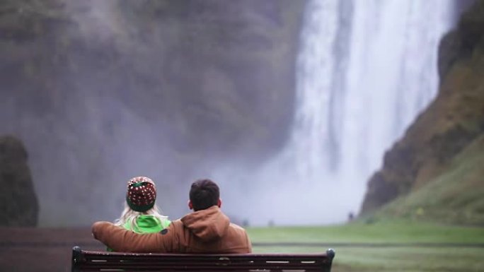 坐在长凳上欣赏冰岛Skogafoss瀑布美景的年轻夫妇的后景