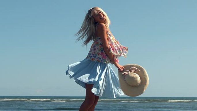 迷人的金发女郎拿着草帽在海滩上旋转