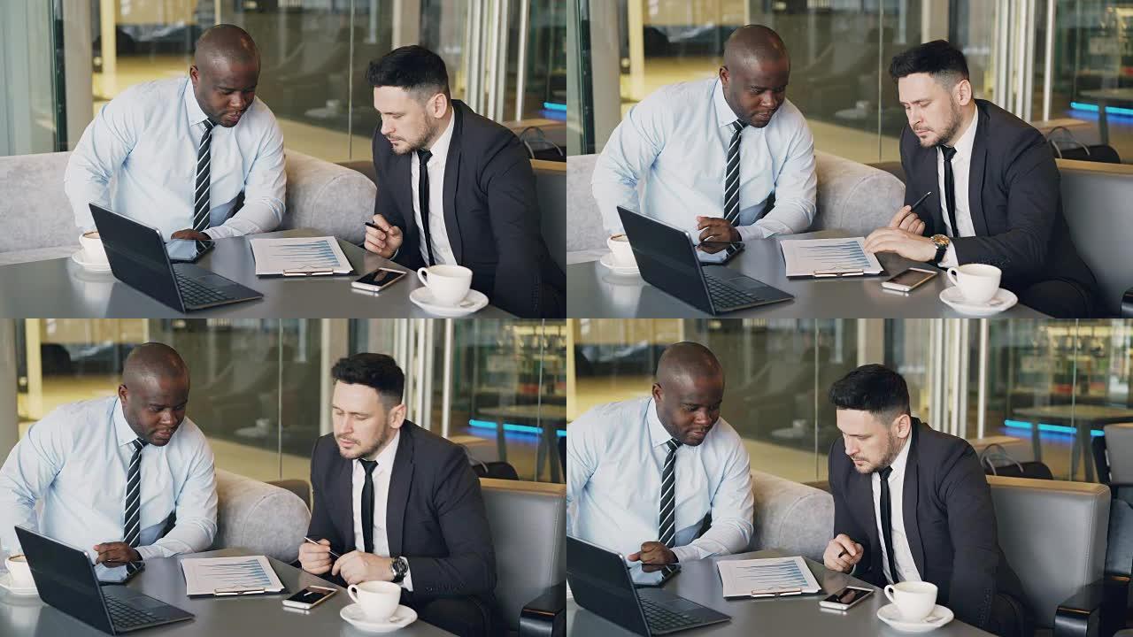 午餐时间，两名穿着正式服装的多种族商业同事坐在餐桌旁讨论财务图表，同时在现代咖啡馆看剪贴板
