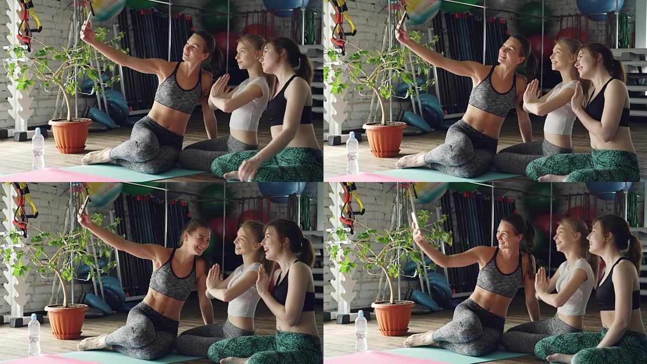 漂亮女孩正在瑜伽馆使用智能手机自拍。年轻女性在聊天，摆姿势和大笑，练习后她们很开心。