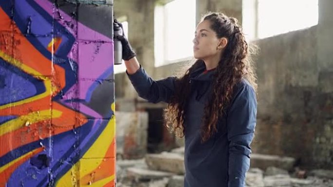 严肃的年轻女性涂鸦艺术家正在使用喷漆在旧仓库的柱子上绘画。背景是废弃的肮脏建筑，地板上有垃圾。