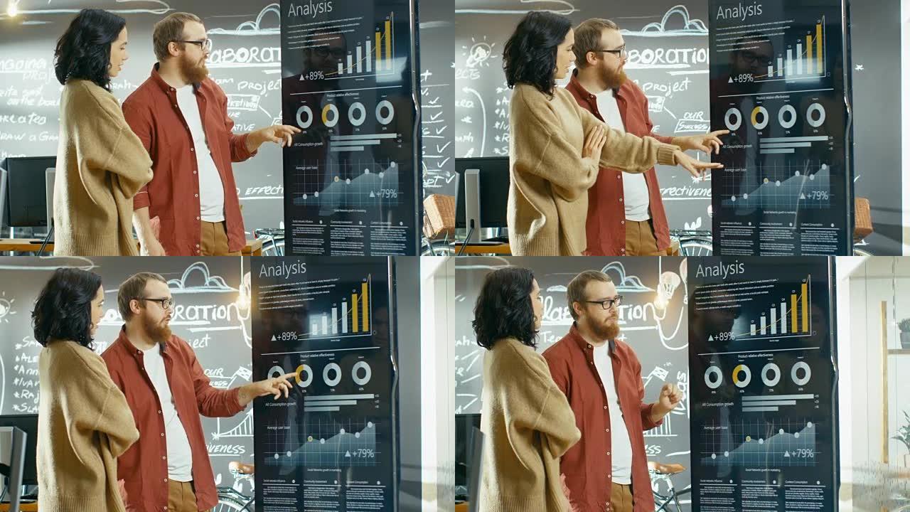 女性开发人员和男性经理使用交互式白板演示触摸屏来查看图表，图形和增长统计数据。他们在时尚的创意工作室