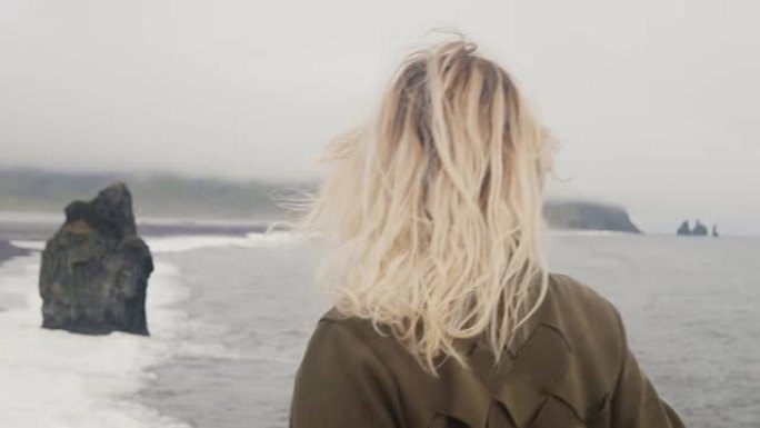 年轻的金发女子站在冰岛的黑色海滩上，看着巨魔脚趾岩石，头发在风中挥舞
