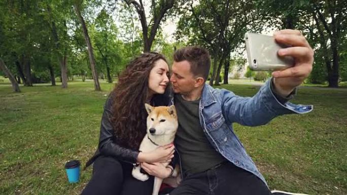 开朗的家伙拿着智能手机自拍，亲吻他的妻子，而她抱着可爱的狗亲吻它。关系、爱情、浪漫和技术概念。