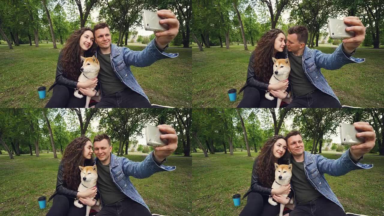 开朗的家伙拿着智能手机自拍，亲吻他的妻子，而她抱着可爱的狗亲吻它。关系、爱情、浪漫和技术概念。