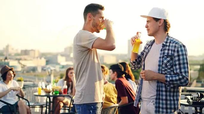 帅哥正在和他的男性朋友聊天，在城市的现代派对上，碰杯和瓶子，站在屋顶上喝酒。年轻人，交流和饮料概念。