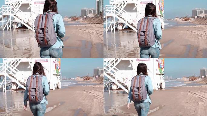 背着背包的女人走在海岸警卫队附近。头发随风飘动。沿着海洋海滩散步的女孩。慢动作