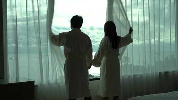 4k镜头的亚洲情侣在浴袍中牵手在一起散步并打开窗帘的场景早晨在豪华酒店的卧室中醒来，生活方式和休闲理