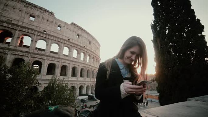 年轻迷人的女人站在意大利罗马的罗马斗兽场附近，使用智能手机。微笑的女孩聊天
