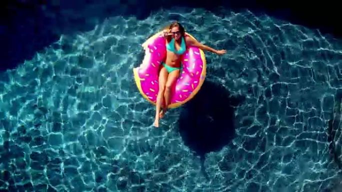 年轻快乐的女人在晴天在蓝色游泳池的充气泳池玩具上放松，放弃相机