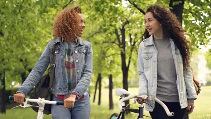 快乐的朋友美丽的女人正拿着自行车在公园里散步，兴高采烈地交谈，背景是美丽的自然树木和草地。友谊和爱好