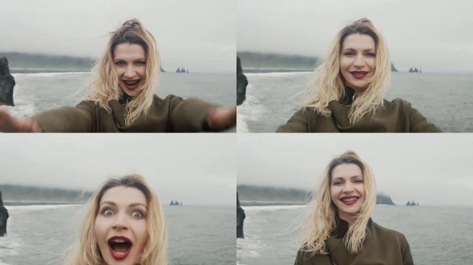 年轻快乐的女人站在黑海滩上，靠近巨魔脚趾岩石，玩得很开心。亲吻相机和微笑