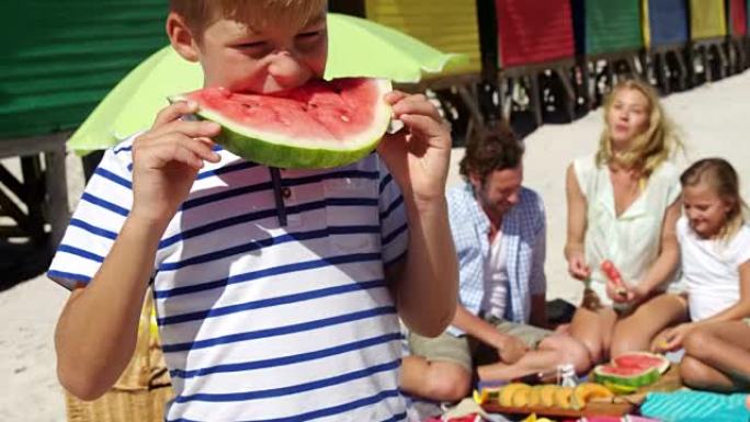 男孩在家人坐在海滩上的背景下吃西瓜