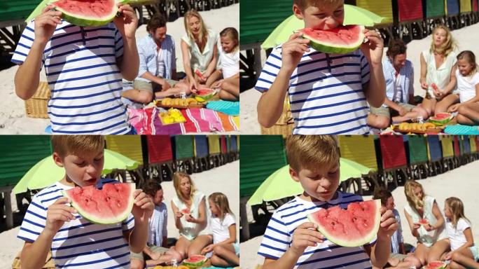 男孩在家人坐在海滩上的背景下吃西瓜