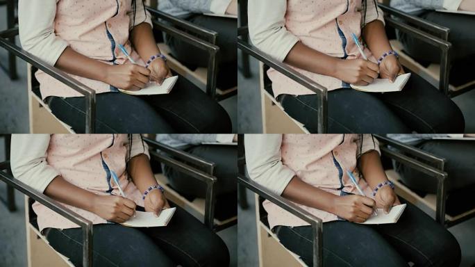 非洲女性手的特写镜头。女人坐在椅子上，用笔记本写字。讲座上的学生女孩