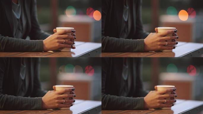 女人的手拿着一杯咖啡的特写镜头，从茶杯里冒烟。模糊的灯光，城市街道