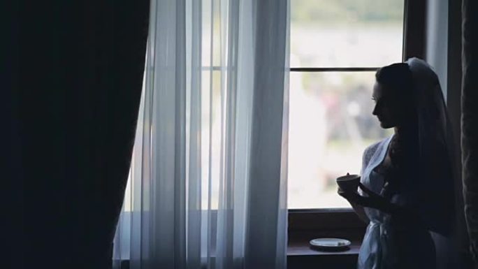 窗户附近黑发新娘的肖像。穿着白色优雅的peignoir的女人喝咖啡，看着距离