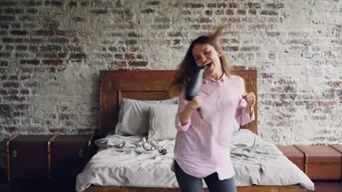 穿着衬衫和牛仔裤的迷人年轻女子拿着吹风机跳舞唱歌，在现代双人床附近的阁楼风格卧室里用它作为麦克风。