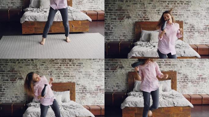 穿着衬衫和牛仔裤的迷人年轻女子拿着吹风机跳舞唱歌，在现代双人床附近的阁楼风格卧室里用它作为麦克风。