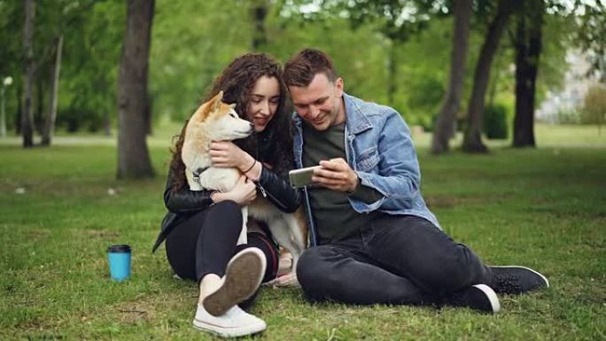 充满爱心的男朋友正在向漂亮的年轻女士展示智能手机屏幕，她坐在城市公园的草坪上抱着并拍拍她的良种狗。人