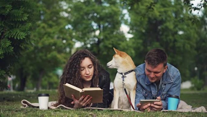 幸福的夫妻在公园休息，男人在使用智能手机，而女人在读书，他们的狗坐在毯子之间。人们在说话和微笑。