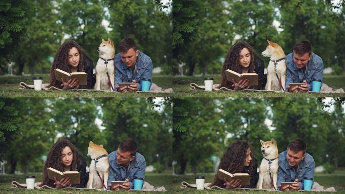 幸福的夫妻在公园休息，男人在使用智能手机，而女人在读书，他们的狗坐在毯子之间。人们在说话和微笑。