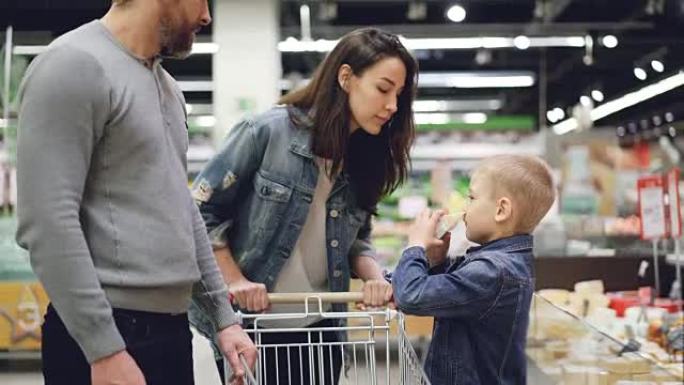 小男孩正在和他的父母一起购物，从冰箱里拿奶酪闻起来，然后送给父母，他们正在检查产品质量并放入购物车。