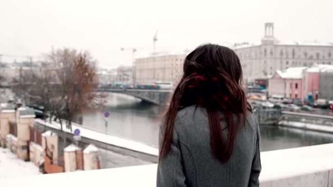 后视图。站在桥上的悲伤的黑发女孩叹了口气，看着冰雪覆盖的冬季小镇