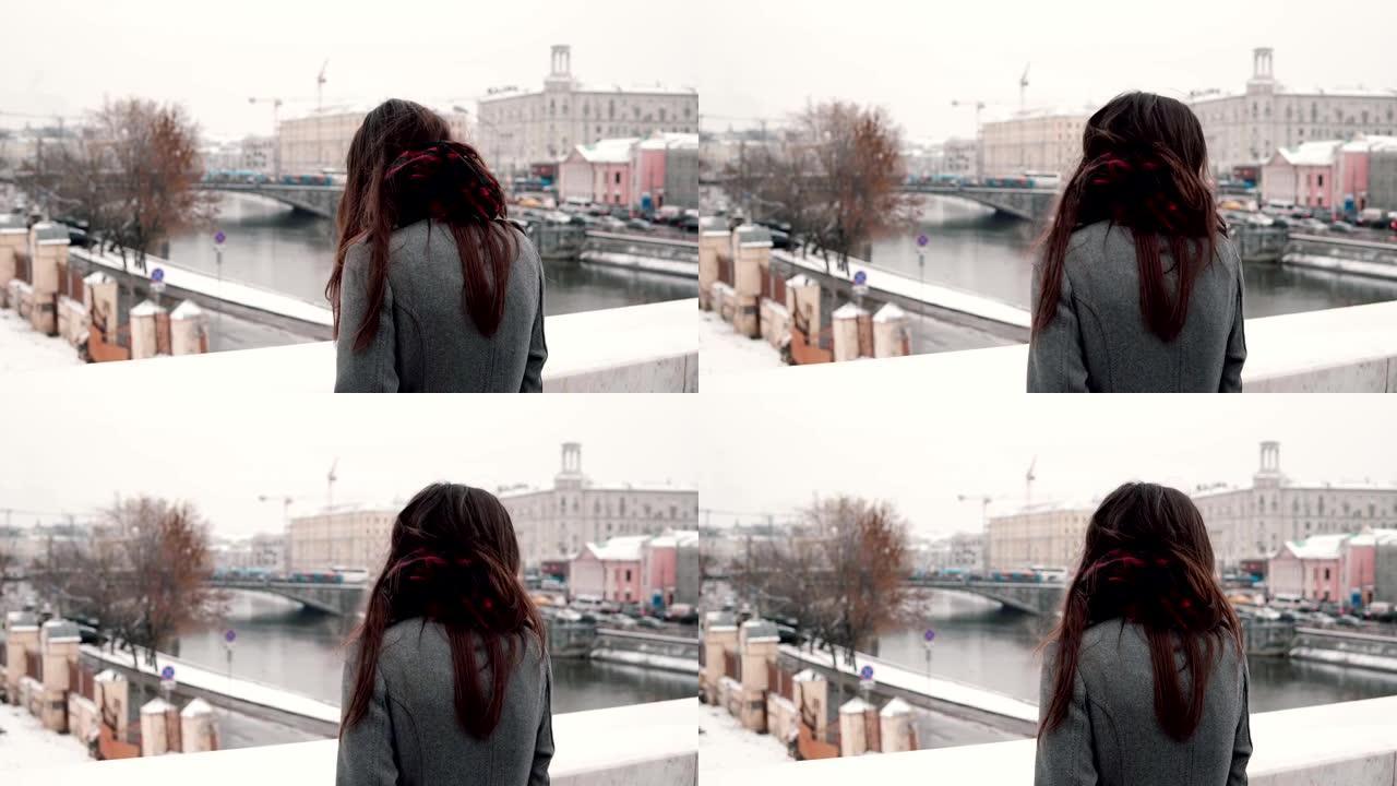 后视图。站在桥上的悲伤的黑发女孩叹了口气，看着冰雪覆盖的冬季小镇
