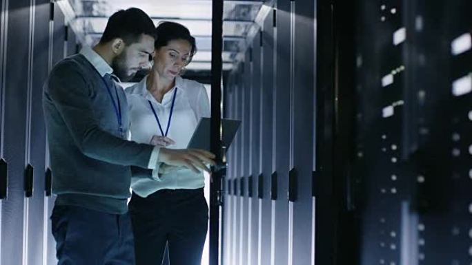 男性IT专家拿着笔记本电脑，与女性服务器技术人员讨论工作。他们站在数据中心，机架服务器柜是开放的。