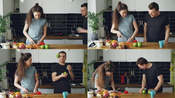 开朗的女人在厨房准备早餐，她的丈夫来拿苹果并展示她的杂耍，女人笑着，一个苹果掉下来，一对夫妇在寻找苹