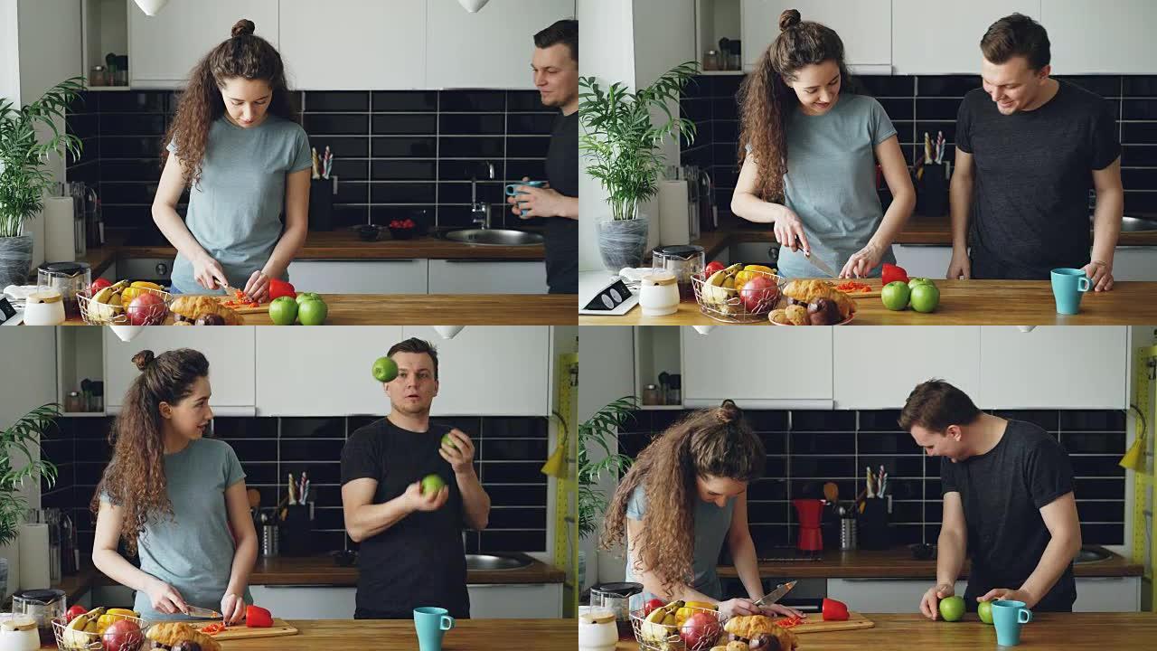 开朗的女人在厨房准备早餐，她的丈夫来拿苹果并展示她的杂耍，女人笑着，一个苹果掉下来，一对夫妇在寻找苹