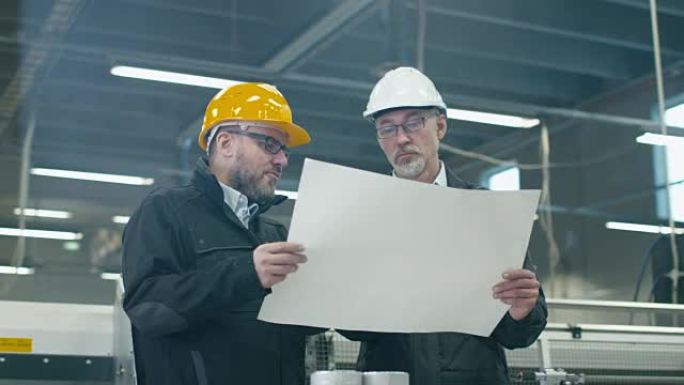 两名戴着安全帽的工程师站在工厂里讨论蓝图。