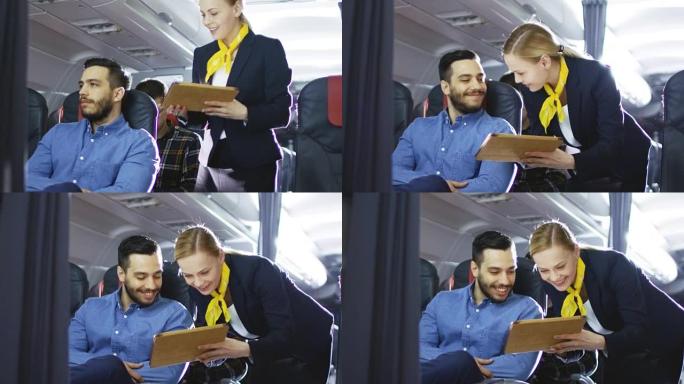 空姐/空姐向西班牙裔男性乘客展示带菜单的平板电脑。他们在机上。商业航空内饰的商务舱是可见的。