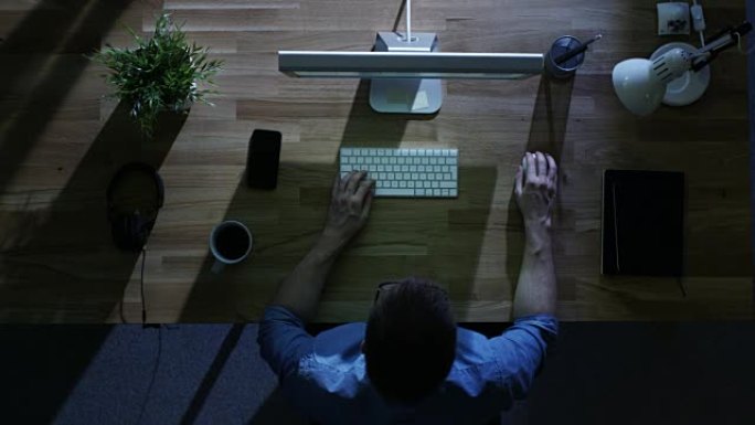 年轻学生在夜间使用模拟绿屏的台式计算机工作的俯视图。他的桌子被外面的冷蓝光照亮。