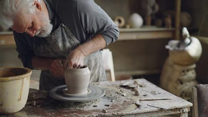 经验丰富的陶艺家白发大胡子的男人正在用湿海绵打磨模制陶瓷锅。可以看到旋转的抛轮，泥泞的工作台和粘土器