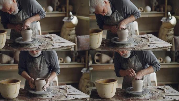 经验丰富的陶艺家白发大胡子的男人正在用湿海绵打磨模制陶瓷锅。可以看到旋转的抛轮，泥泞的工作台和粘土器