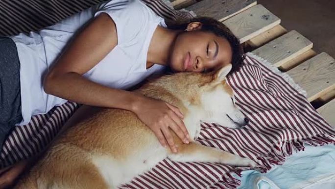 美丽的非洲裔美国少年和可爱的宠物狗一起睡在木床上，女孩穿着舒适的睡衣和拥抱动物。