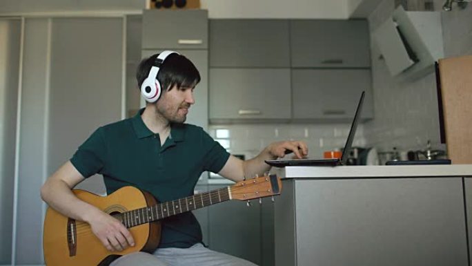 开朗的年轻人戴着耳机坐在厨房里学习在家中使用笔记本电脑弹吉他