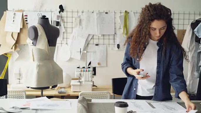 服装设计师enterpreneur正在使用智能手机在工作室的桌子上拍摄服装素描和咖啡的平铺。年轻迷人