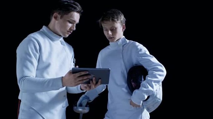两名装备齐全的年轻击剑手使用平板电脑了解更多关于击剑中的战略、进攻和防守的知识。拍摄孤立在黑色背景上